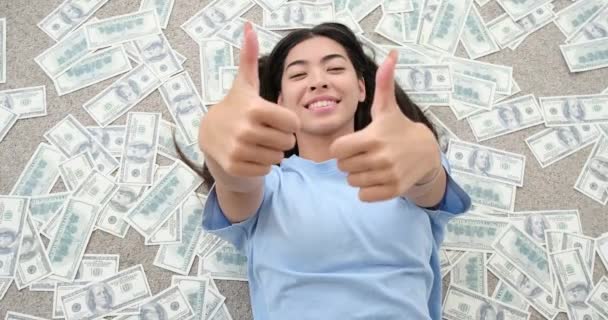 Kvinnen som gir tomler opp gester mens hun ligger på spredte dollarsedler – stockvideo