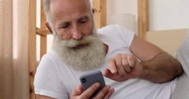 Yaşlı adam yatakta cep telefonu kullanıyor.