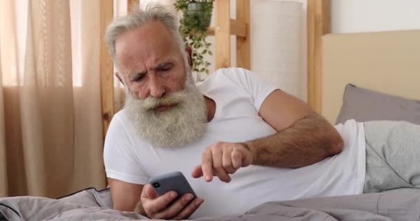 Hombre viejo frustrado usando el teléfono móvil en la cama — Vídeos de Stock