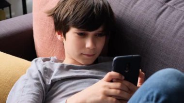 Kanepede uzanan ve parlak bir oturma odasında akıllı telefon kullanan bir gencin yan görüntüsü..