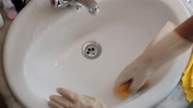 Yetişkin bir kadının sabunlu banyo lavabosunu süngerle temizlerken, eldiven giyerken, yüzü görünmezken tepeden bakışı..