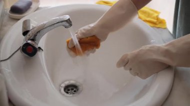 Yetişkin bir kadının banyo lavabosunda eldiven takıp, sarı bir mikrofiber bezi duruladığı ve sıktığı yakın plan..