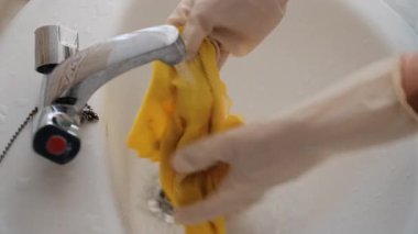 Yetişkin bir kadının banyo lavabosunda eldiven giyerken mikrofiber temizleme bezini duruladığı ve sıktığı yakın plan..