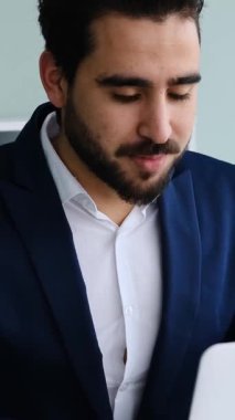 Yetişkin adam modern bir ofis ortamında dizüstü bilgisayar ve belgelerle yumuşak doğal ışık altında çalışır. Yavaş çekimde yakalanır..