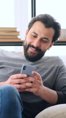 Yetişkin bir adam evdeki kanepede dinlenir, kaydırırken gülümser ve akıllı telefonuyla kitap okur..