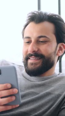 Yetişkin bir adamın yavaş çekimde kanepeye yaslanıp akıllı telefon kullanırken gülümsemesi..