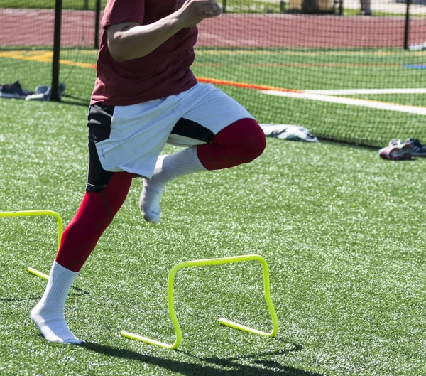 A high school track and field athlete is performing a speed and agility drill over yellow mini banana hurdles on a green turf field with no shoes on.