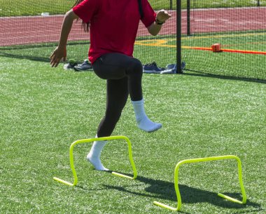 Bir lise kadın atlet formu matkaplar üzerinde ayakkabı ile steping tarafından yeşil çim alan sarı mini engellerin üzerinden gerçekleştiriyor.