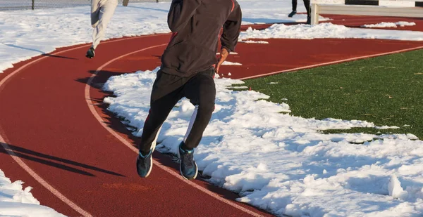 Boys running fast in lane one of a red track with snow on both sides of them during winter track and field practice.