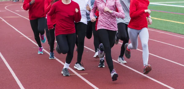 A high school cross country and track teams run together in a group on a red track during practice.