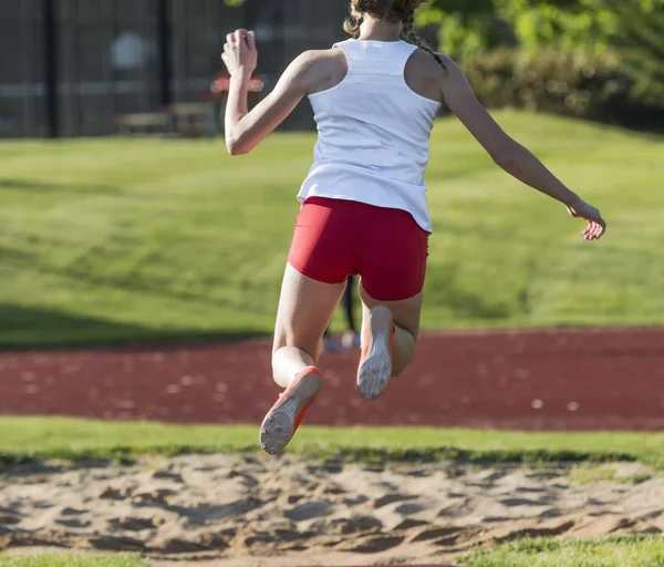 View from behind of a teenage girl triple jumping at a track and field competition, flying in the air toward the sand pit.