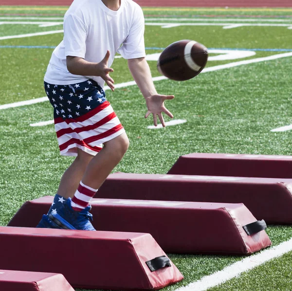 A young boy is at summer football camp catching a football while running sideways over red barriers and wearing red, white and blue flag shorts and socks.
