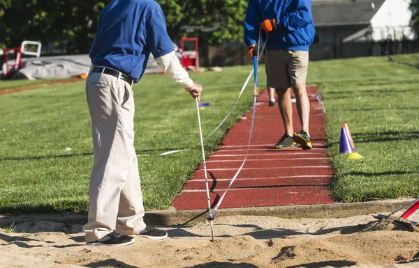 Two track and field officials are measuring a long or triple jump distance at a competition outdoors on a sunny day.