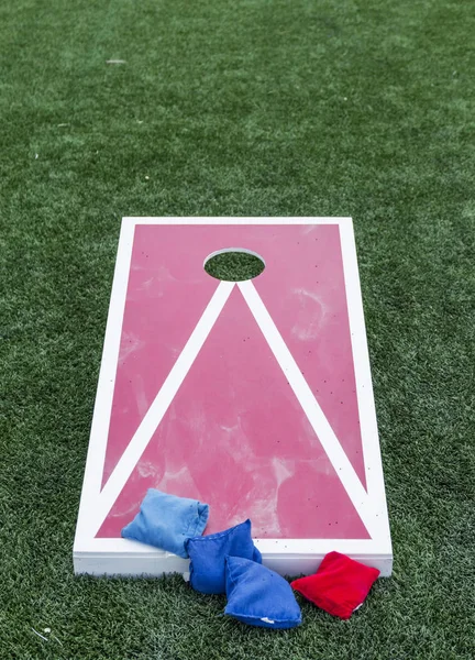 A wood red and white corn hole board is set up on a green turf field with blue and red bean bags.