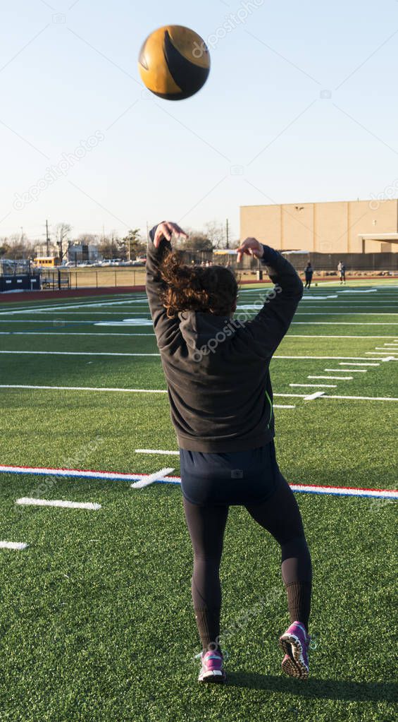 An athlete is on a green turf field throwing a black and gold medicine ball over her head backwords during track and field practice.