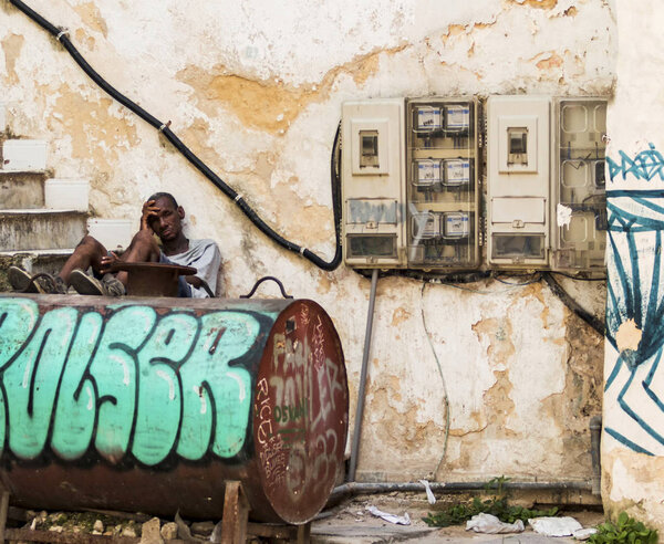 Гавана, Куба - 25 июля 2018 года: Кубинский мужчина, сидящий на ступеньках с ногами на нефтяном танке, прислонившись к зданию в тени, избегая жары
.