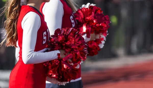 Two high school cheerleaders with red and white pompoms are watching the football game ready to cheer for their team.