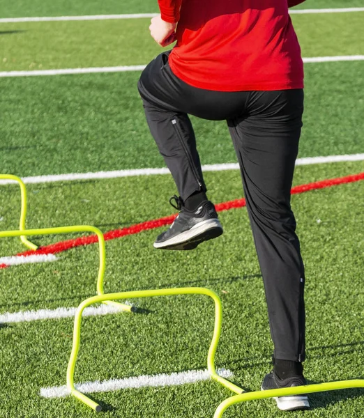 A high school athlete is performing speed and agility sports training drills stepping over yellow mini hurdles on a green turf field.
