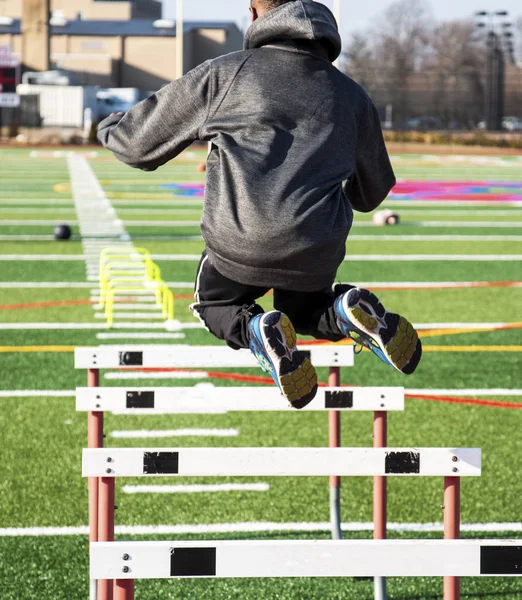 An athlete is jumping over track hurdles on a green turf field with medicine balls in the background during speed and strength practice.
