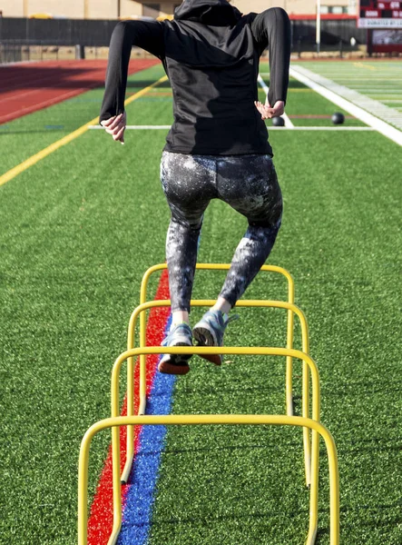 A high school female track and field athlete is jumping over two feet high yellow mini hurdles on a green turf field.