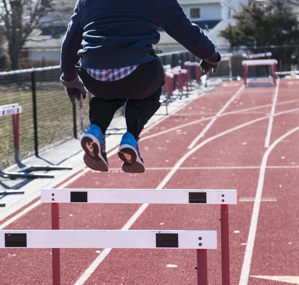 A track and field runner is jumping over hurdles on a red track during winter with gloves on.