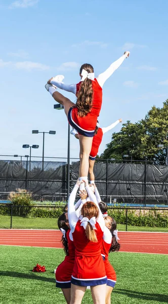 A pyramid of high school cheerleaders are holding their teammates in the air by their ankles practicing a routine on green turf.