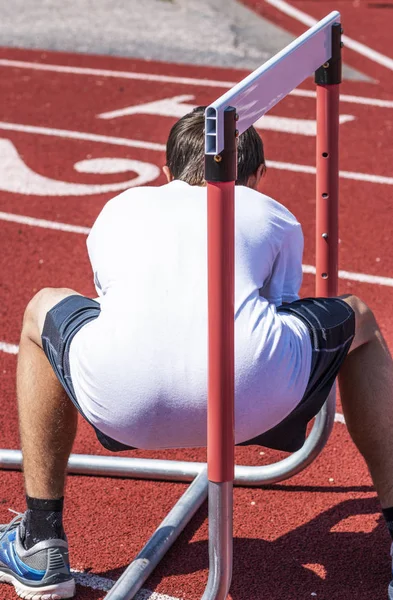 A young athlete is squatting underneath a track hurdle during strength and agility practice.