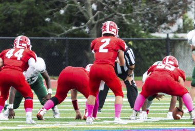 A high school football quarterback in a red uniform is in the shotgun ready to receive the ball to start the play.