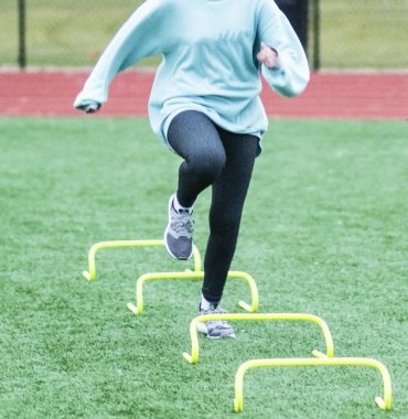 A female high school athlete is training for speed and agility by running over yellow mini hurdles, also known as banana hurdles, on a green turf field. clipart