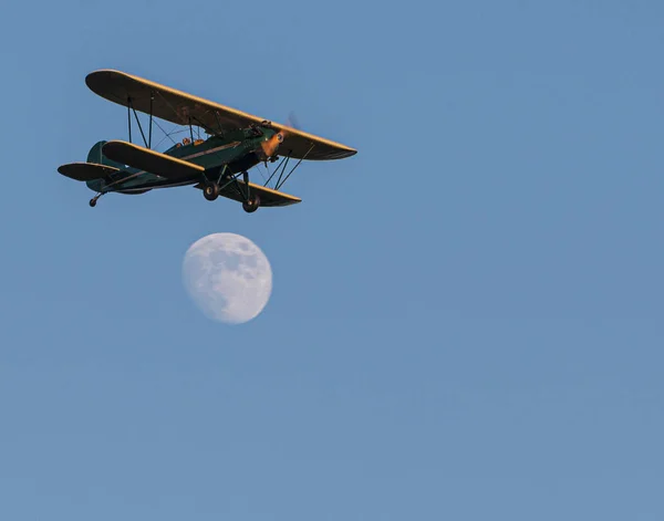 Green and yellow bi-plane flying over an almost full moon