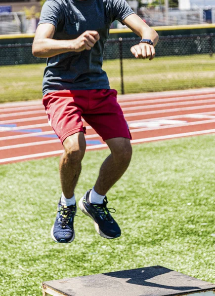 Jovem atleta fazendo caixa salta em um campo de relva com pista nas costas — Fotografia de Stock