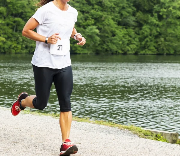 A women in a white shirt and black spandex running a 10K race in Babylon Village passing Southards Pond in the summer.
