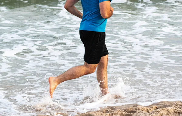 Side View of a man running and splashing in the water on the edge of the beach barefoot.