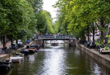 Amsterdam, Hollanda - 5 Ağustos 2025: Ağaçlar ve teknelerle dolu, büyüleyici bir köprüye sahip sakin bir kanal.