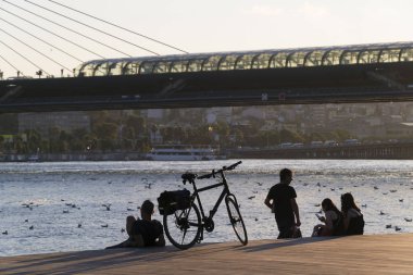 İstanbul, Türkiye - 19 Temmuz 2019 : Bir motorcu, bisikleti bir erkek ve iki genç kadın gün batımında ahşap zeminüzerinde Haliç sahilinde. İstasyonlu Metro Köprüsü ve arka planda şehir manzarası olan Unkapanı Köprüsü vardır..