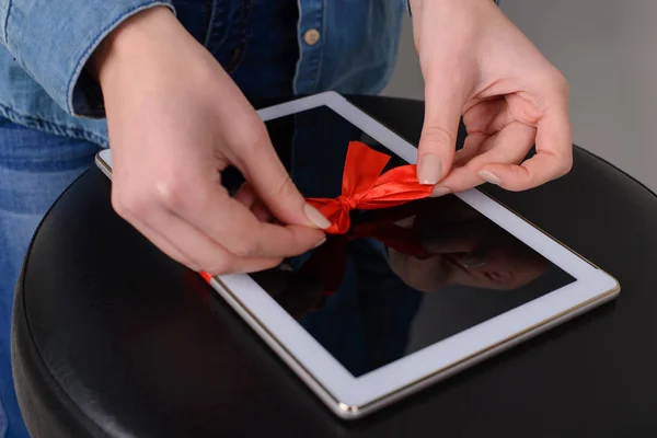 Woman's hands tie digital white tablet with red ribbon. She prepares for winter holidays