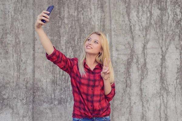 Woman in casual clothes with beaming smile taling selfie on her smartphone and showing v-sign