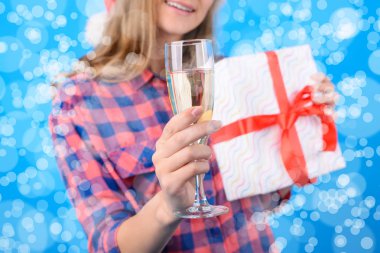 Bir cam og şampanya kadının elinde fotoğraf kırpılmış, üzerinde mavi arka plan izole bir hediye kutusu tutuyor