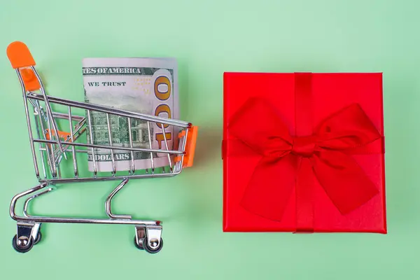E-commerce concept last chance sale birthday. Top above high angle flat lay close up photo of bright ribbon little package and trolley with 100 dollars isolated on pastel green background