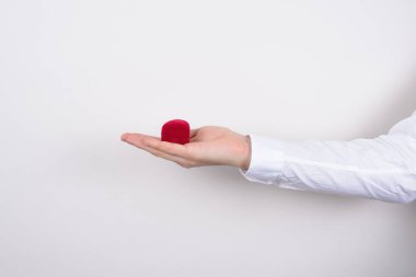 Side profile closeup cropped photo of hand holding closed red heart shaped small little box with ring inside isolated grey background