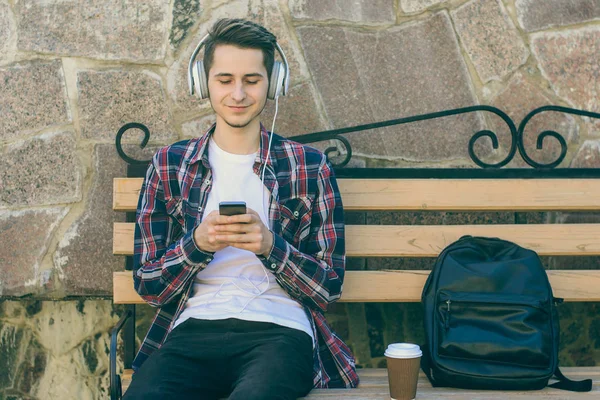 Young smiling guy sitting on a bench and using his smartphone fo — Stock Photo, Image