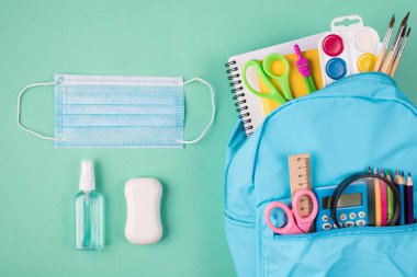 Sağlık ve öğrenim konsepti. Üstte mavi sırt çantasının renkli maske sabunu ve el dezenfektanı ile dolu resmi turkuaz arka planda izole edilmiş.