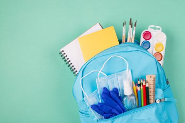 Okul konseptinde sağlık hizmeti. Üstünde renkli kağıtlarla dolu sırt çantasının resmi var. İki mavi maske lastik eldiven ve el dezenfektanı turkuaz arka planda izole edilmiş.