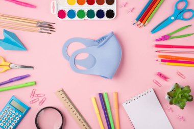Uzaktan çalışma konsepti. Quarantime konsepti. Yukarıda mavi maskenin yukarıdan çekilmiş fotoğrafı. Etrafı renkli kırtasiyelerle çevrili. Pastel pembe arka planda izole edilmiş.