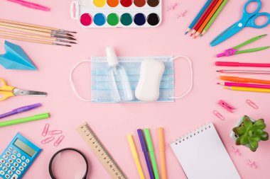 Coronavirus önleme konsepti. Üzerinde mavi maske sabun el dezenfektanı ve pastel pembe arka planda izole edilmiş sulu renkli kırtasiye malzemesi resmi var.