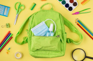 Karantina sırasında sağlık sigortası. Üstünde yeşil sırt çantası maskeli el dezenfektanı ve sarı arka planda izole edilmiş renkli kırtasiye malzemesi resmi var.