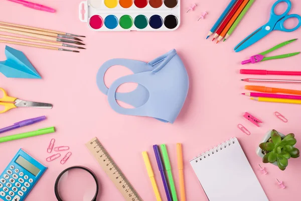 Uzaktan çalışma konsepti. Quarantime konsepti. Yukarıda mavi maskenin yukarıdan çekilmiş fotoğrafı. Etrafı renkli kırtasiyelerle çevrili. Pastel pembe arka planda izole edilmiş.