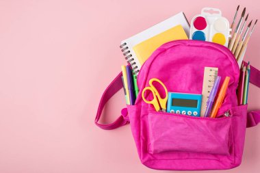 Üst tepede pembe sırt çantasının ve sağ tarafa yerleştirilmiş renkli kırtasiyenin fotosu var. Kopyalama alanı olan pastel pembe arka planda izole edilmiş.