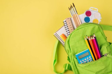 Tepede yeşil sırt çantasının, telif alanı olan sarı arkaplanda izole edilmiş renkli kırtasiyeli resmi var.