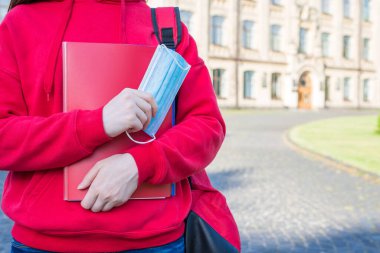 Tıbbi maske takmak ve sosyal mesafeyi korumak gerekli. Kırmızı tişörtlü, elinde kitaplarla yüz maskesi olan genç bir kızın yakın plan fotoğrafı.
